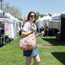 Load image into Gallery viewer, A smiling woman at outdoor market with Bumkins Packable Tote Bag: Yoga Pals on her shoulder holds a snack, wearing sunglasses and denim dress.