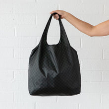 Load image into Gallery viewer, A hand holds a Bumkins Packable Tote Bag: Rich Black against a white brick wall.