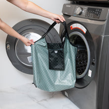 Load image into Gallery viewer, A person holds a Bumkins Packable Tote Bag: Rich Black in front of an open front-loading washing machine.