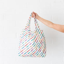 Load image into Gallery viewer, A hand holds a Bumkins Packable Tote Bag: Mickey & Friends Varsity against a white brick wall.