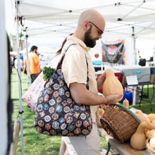 Load image into Gallery viewer, A man with a Bumkins Packable Tote Bag: Ghostly Gallery over his shoulder examines a butternut squash at an outdoor market under a white tent.