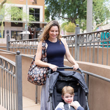 Load image into Gallery viewer, A woman smiles as she carries a Bumkins Packable Tote Bag: Ghostly Gallery and pushes a stroller outdoors, surrounded by trees and railings.