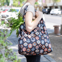 Load image into Gallery viewer, A person carries a Bumkins Packable Tote Bag: Ghostly Gallery, filled with green veggies on a bustling city street.