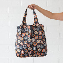 Load image into Gallery viewer, A hand holds a Bumkins Packable Tote Bag: Ghostly Gallery against a white brick wall.