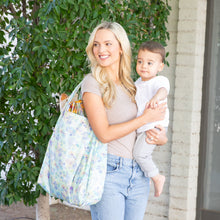 Load image into Gallery viewer, A woman smiles, carrying a child and a Bumkins Packable Tote Bag: Aloha Stitch near lush foliage outdoors.