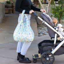 Load image into Gallery viewer, A person strolls outdoors with a baby in a stroller, carrying a Bumkins Packable Tote Bag: Aloha Stitch over their arm.
