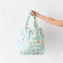 Load image into Gallery viewer, A hand holds a Bumkins Packable Tote Bag: Aloha Stitch against a white brick wall.
