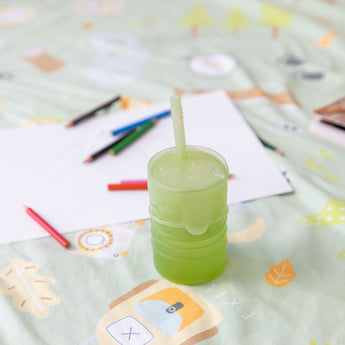 Bumkins Silicone Straw Cup with Lid: Sage sits on a patterned blanket among paper and colored pencils.