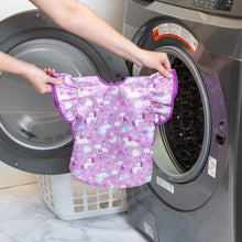 Load image into Gallery viewer, A person holds Bumkins Ruffle Apron Bib: Unicorns in front of an open front-loading washing machine.