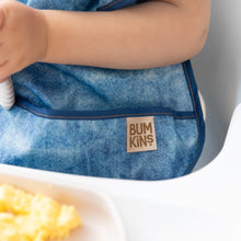 Load image into Gallery viewer, Close-up of a child in a high chair wearing a Bumkins Ruffle Apron Bib: Crushed Denim, with focus on the pocket.