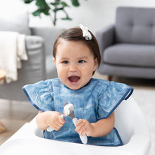 Load image into Gallery viewer, A girl in a blue bib beams in a high chair, holding Bumkins Spoon + Fork: Sand in a cozy living room.