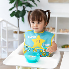 Load image into Gallery viewer, A girl with pigtails in a high chair wears a Bumkins SuperBib® 2 Pack: Super Mario™ Expressions, holding a spoon near a blue bowl.