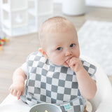 A baby in a Bumkins SuperBib® 3 Pack: Palms + Check sits in a high chair, nibbling curiously with a bowl in front of them.