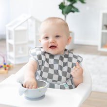 Load image into Gallery viewer, A baby in a high chair, smiling in a Bumkins SuperBib® 3 Pack: Palms + Check, enjoys eating from a bowl with a small playhouse nearby.