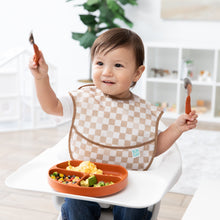 Load image into Gallery viewer, A smiling toddler in a Bumkins SuperBib® 3 Pack: Palms + Check sits in a high chair, holding utensils with food in a divided plate on the tray.