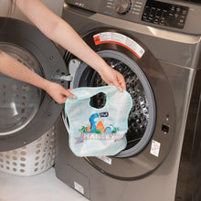 Load image into Gallery viewer, A person holds a Bumkins SuperBib® 3 Pack: Hangry, Dinosaurs, Blue Tropic in front of an open front-loading washing machine.