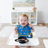 A smiling baby in a high chair, wearing a blue Bumkins SuperBib®: Mickey & Friends MVP, holds a spoon while eating cereal in a bowl.