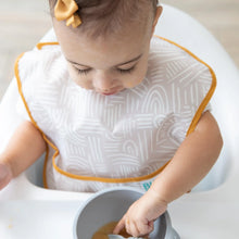 Load image into Gallery viewer, A toddler in a high chair uses a spoon to eat from a bowl, wearing a beige Bumkins SuperBib® 3 Pack: Desert Boho.