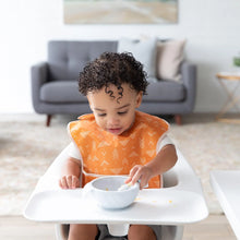 Load image into Gallery viewer, A toddler in an orange Bumkins SuperBib® 3 Pack: Desert Boho eats happily in a high chair, with a cozy living room in the background.