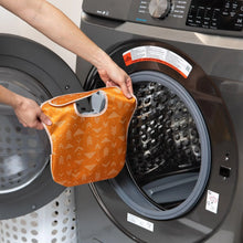 Load image into Gallery viewer, A person holds an orange Bumkins SuperBib® 3 Pack: Desert Boho in front of an open washing machine.