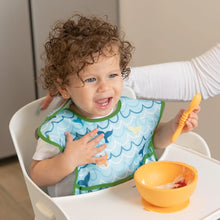 Load image into Gallery viewer, A curly-haired toddler in a high chair wears a Bumkins SuperBib® 3 Pack: Rolling With the Waves, holding a spoon & smiling by an orange bowl.