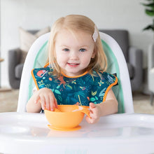 Load image into Gallery viewer, A toddler sits in a high chair, wearing a Bumkins SuperBib® 3 Pack: All Together Now while holding an orange bowl.