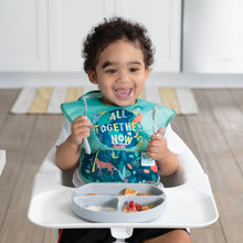 Load image into Gallery viewer, A smiling toddler in a high chair wears a Bumkins SuperBib® 3 Pack: All Together Now, holding utensils near a gray divided plate with food.