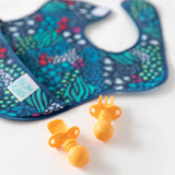 A Bumkins SuperBib® 3 Pack: All Together Now lays out of focus on a white background, with orange silicone utensils in the foreground.