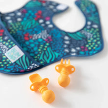 Load image into Gallery viewer, A Bumkins SuperBib® 3 Pack: All Together Now lays out of focus on a white background, with orange silicone utensils in the foreground.