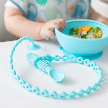 Load image into Gallery viewer, Close-up of a baby in a bib holding a bowl, with a blue spoon attached to a Bumkins Silicone Accessory Tether 2-Pack: Blue at a white table.