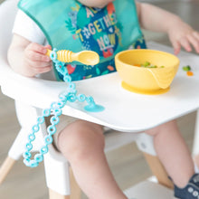 Load image into Gallery viewer, A baby explores a yellow bowl of food on a high chair tray, holding a spoon attached with a Bumkins Silicone Accessory Tether 2-Pack: Blue.