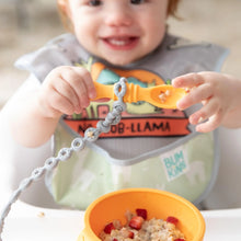 Load image into Gallery viewer, A child holds a spoon attached to a Bumkins Silicone Accessory Tether 2-Pack: Gray over a bowl of oatmeal on a high chair tray.