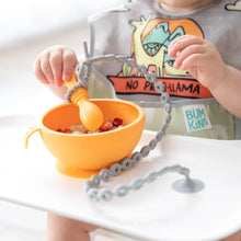 Load image into Gallery viewer, A baby in a high chair eats from an orange bowl, with a spoon attached to the tray with a Bumkins Silicone Accessory Tether 2-Pack: Gray.