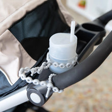 Load image into Gallery viewer, A gray cup in a stroller's cup holder is secured to the handle by a Bumkins Silicone Accessory Tether 2-Pack: Gray.