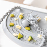 Close-up of a gray toddler fork attached to a Bumkins Silicone Accessory Tether 2-Pack: Gray on a gray placemat with avocado pieces.