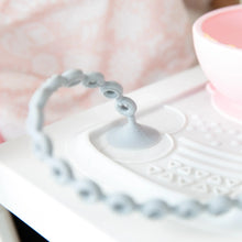 Load image into Gallery viewer, Close-up of a Bumkins Silicone Accessory Tether 2-Pack: Gray on a high chair tray, with the suction cup attached to a placemat.
