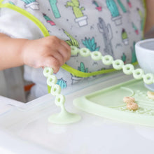 Load image into Gallery viewer, Close-up of a baby wearing a cactus-print bib in a high chair holds a Bumkins Silicone Accessory Tether 2-Pack: Sage attached to the tray.