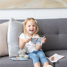 Load image into Gallery viewer, A smiling young girl on a sofa holds Bumkins Reusable Snack Bag, 3-Pack: Princess Magic, Ariel, and Jasmine, a stuffed animal beside her.
