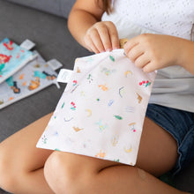 Load image into Gallery viewer, A child on couch opens a Bumkins Reusable Snack Bag, 3-Pack: Princess Magic, Ariel, and Jasmine, with two bags beside her.