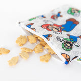 Close-up of bunny-shaped crackers spilling from a Bumkins Reusable Snack Bag, Large: Super Mario™ & Luigi on a white background.
