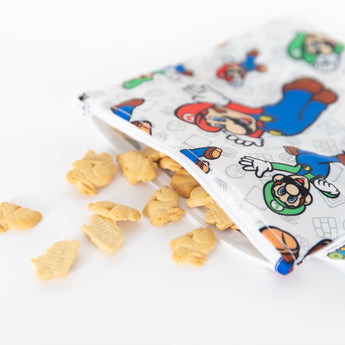 Close-up of bunny-shaped crackers spilling from a Bumkins Reusable Snack Bag, Large: Super Mario™ & Luigi on a white background.