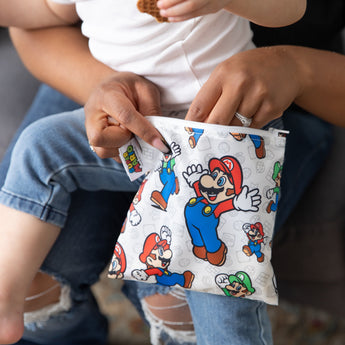 A person reaches into a Bumkins Reusable Snack Bag, Large: Super Mario™ & Luigi while a baby sits on their lap.