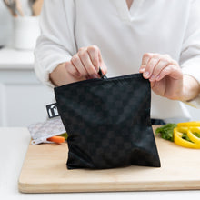 Load image into Gallery viewer, A person zips a Roamio by Bumkins Reusable Snack Bag, Large 2 Pack: Rich & Boujee on a cutting board with veggie slices behind.