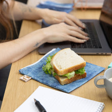 Load image into Gallery viewer, A sandwich sits on top of a Roamio by Bumkins Reusable Snack Bag, Large 2 Pack: Denim on Denim on a desk next to a person using a laptop.