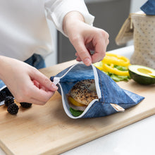 Load image into Gallery viewer, A person opens a Roamio by Bumkins Reusable Snack Bag, Large 2 Pack: Denim on Denim on a cutting board revealing a sandwich inside.