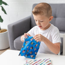 Load image into Gallery viewer, A boy in a white shirt opens Bumkins Reusable Snack Bag, Large 2 Pack: Mickey & Friends MVP at a table in front of a couch and potted plant.
