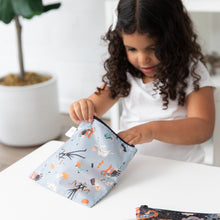 Load image into Gallery viewer, A young girl with curly hair opens a Bumkins Reusable Snack Bag, Large 2 Pack: Everyday Is Halloween at a table, a plant in the background.