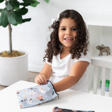 Load image into Gallery viewer, A curly-haired girl at a table reaches into a Bumkins Reusable Snack Bag, Large 2 Pack: Everyday Is Halloween, with a plant and shelves nearby.