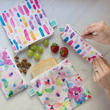 Bumkins Reusable Snack Bag, Large 2-Pack: Watercolor & Brush Strokes lays on a marble surface with fruit and a sandwich; a hand unzips another bag nearby.