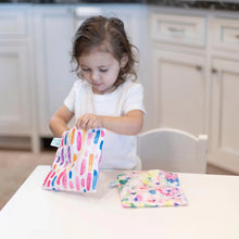 Load image into Gallery viewer, A child joyfully opens a Bumkins Reusable Snack Bag, Large 2-Pack: Watercolor & Brush Strokes, with the other bag beside on a table.
